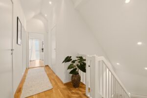 Utsnitt av trappegang/mellomgang på øvre plan. Det er downlights i taket og glassfelt i veggen med sikt til stuen under.